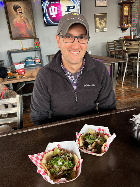 The Grey Eagle Taqueria in Hot Springs, NC with white brunette male in gray hat with glasses sitting with two takeaway soft tortilla tacos in Big Pillow Brewing Taproom