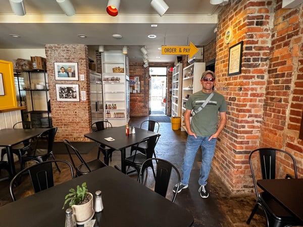Zadie's Market, Bar, and Restaurant in Marshall NC with image of white brunette male in hat, t-shirt, jeans, and sunglasses standing in brick dining room with tables and chairs