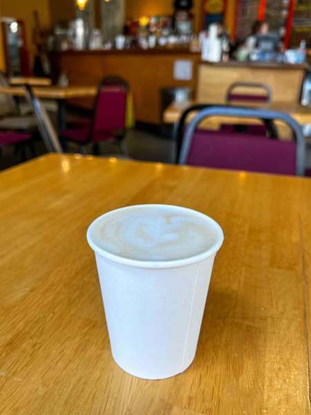 Zuma Coffee and Provisions in Marshall NC with image of takeaway latte on brown table with red chairs in background