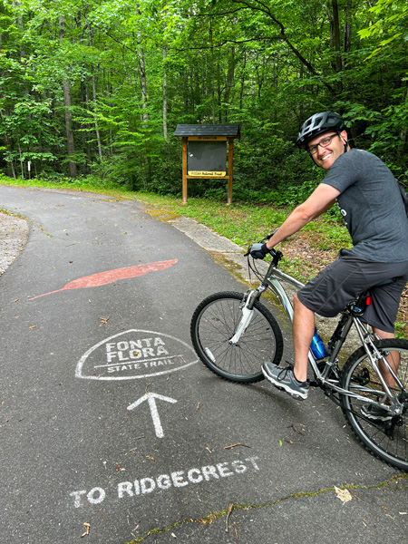 Fonta Flora State Trail Old Fort NC trailhead marker on pavement with red feather and with white brunette male on mountain bike with helmet on paved pathway