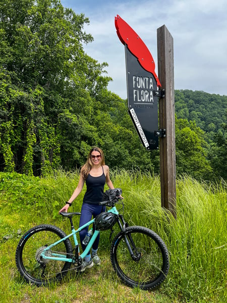 Fonta Flora State Trail Point Lookout in Old Fort NC with sign at top of the point with red feather and white brunette female with deep mint green mountain bike standing in front of it