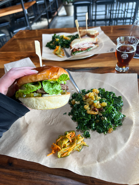 Hillman Beer in Old Fort, NC with white hand holding up burger with lettuce, mayo, mustard, and pickles in front of plate with kale salad and pickle carrot salad