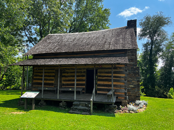 Mountain Gateway Museum in Old Fort North Carolina historic cabin with brown wood, porch and house roof, and chimney on green grass with blue sky and trees in background