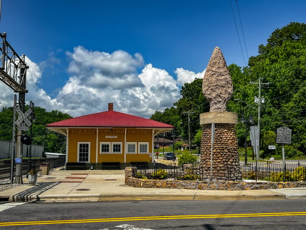 Old Fort Railroad Museum, Train Depot, and Arrowhead Monument with yellow facade and red roof structure next to concrete and stone arrowhead statue with railroad crossing, road, and blue cloudy sky