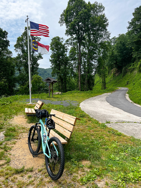 Point Lookout in Old Fort North Carolina with deep mint green bike leaning on a wooden bench with state and American flag in background with informational trail board, green trees, and overcast sky