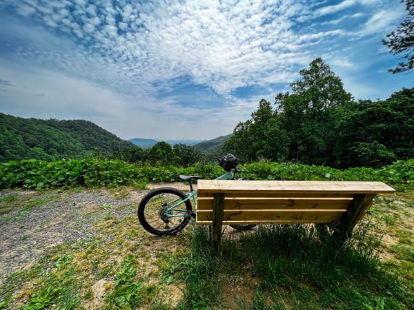 Point Lookout Trail Old Fort NC with wooden bench, bike leaning on bench, and overlook to blue, gray and green mountains with blue cloudy sky