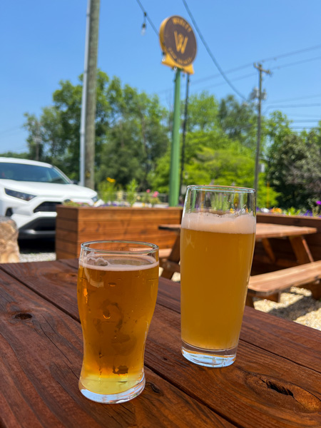 Whaley Farm Brewery Old Fort NC with yellow and orange brown beer on picnic table outside with sign for brewery blurred in background against blue sky