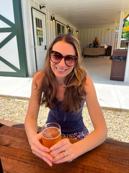 Whaley Farm Brewery Old Fort NC image of Christine, a white female with brunette hair with golden highlights wearing sunglasses and holding orange beer at picnic table outside
