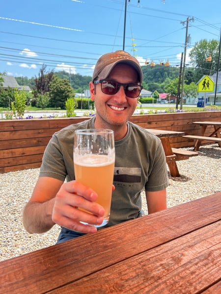 Whaley Farm Brewery Old Fort NC with white male in green t-shirt and ssunglasses holding up light yellow gluten-reduced beer at picnic table outside