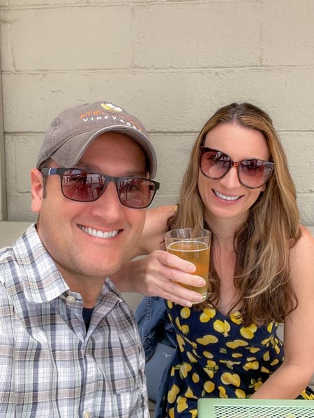 Eluvium Brewing Company Weaverville with white brunette and blonde highlights woman with sunglasses in lemon-printed dress holding up a Helles Beer next to white brunette male with hat and sunglasses in plaid striped shirt