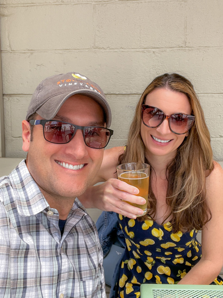 Eluvium Brewing Company in Weaverville with white brunette and blonde highlights woman with sunglasses in lemon-printed dress holding up a Helles Beer next to white brunette male with hat and sunglasses in plaid striped shirt