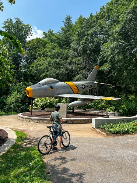 F 86 Sabre plane Memorial in Cleveland Park in Greenville SC with white male on mountain bike with helmet stopped in front of the gray plane with yellow stripe