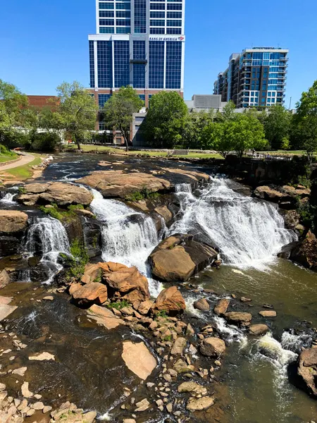 Falls Park on the Reedy in Greenville, SC view from Liberty Bridge with waterfalls going over rocks with city buildings in the background
