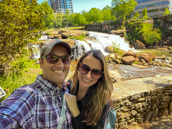 Falls Park on the Reedy selfie on Liberty Bridge in Greenville SC with white brunette male and female wearing sunglasses and falls and cityscape in the background