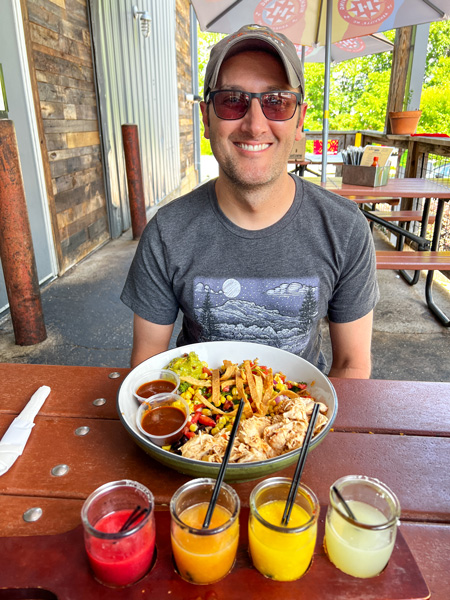 Tom, a white brunette male with sunglasses in a t-shirt, sitting with a bowl of food and brightly-colored margarita flight at Farmhouse Tacos Restaurant in Travelers Rest, SC 