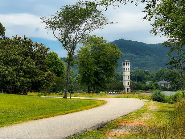 Furman University clock tower and paved pathway around Furman Lake along the Swamp Rabbit Trail in Greenville SC; you can see green grass, a blue cloudy sky, and mountains in the background