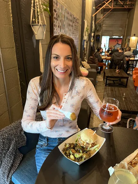 Ginger's Revenge in Asheville NC with white brunette woman in pink top and jeans with Gluten Free Beer in hand and eating a vegan and gluten-free burger with Brussels sprouts
