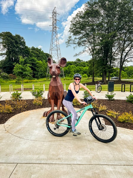 Greenville SC Swamp Rabbit Trail Guide with white female wearing helmet, sunglasses, purple and gray leggings, and black top on deep mint mountain bike in front of brown rabbit sculpture that is slightly larger than her