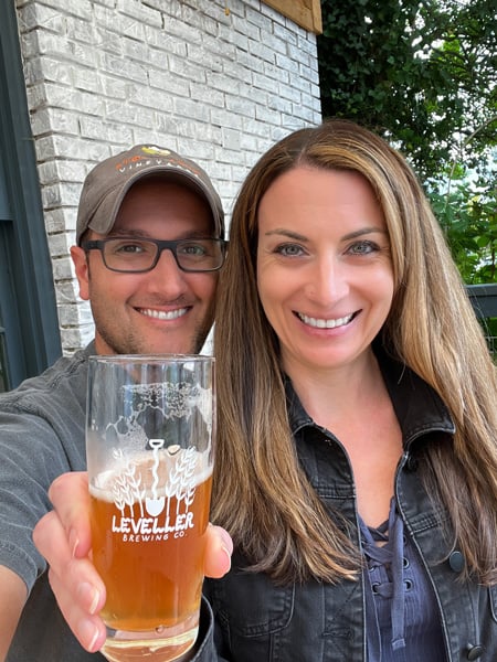 Tom and Christine, a white brunette male with hat and glasses and a white brunette female with blonde highlights, holding up an orange brown beer in a Leveller glass outside on the patio at Leveller Brewing Co in Weaverville, North Carolina