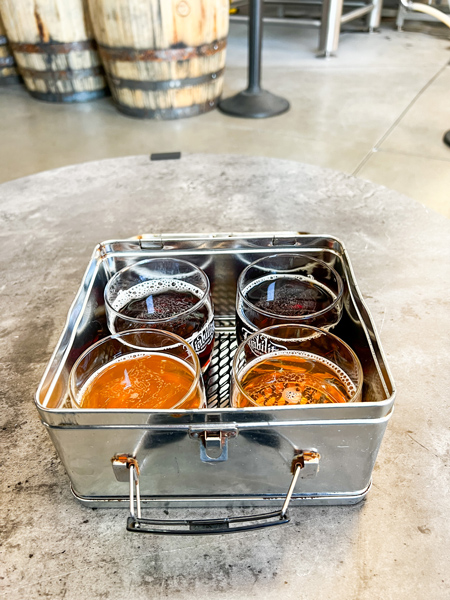 Liability Brewing Co Gluten Reduced Beer in Greenville SC with flight of two orange and two brown beers in a tin on a table with barrels blurred in background