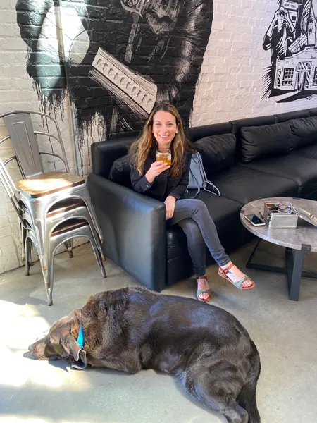 Liability Brewing Co in Greenville SC Gluten Reduced Brewery with Christine, a white brunette female with blonde highlights in jeans and black jean jacket, holding an orange beer with a large black dog at her feet and mural on the wall behind her