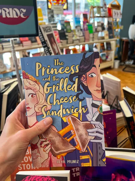 Malaprop's Bookstore Book Shopping in Asheville NC with white hand holding up a graphic novel called The Princess and the Grilled Cheese with image of two people splitting a cheesy grilled cheese