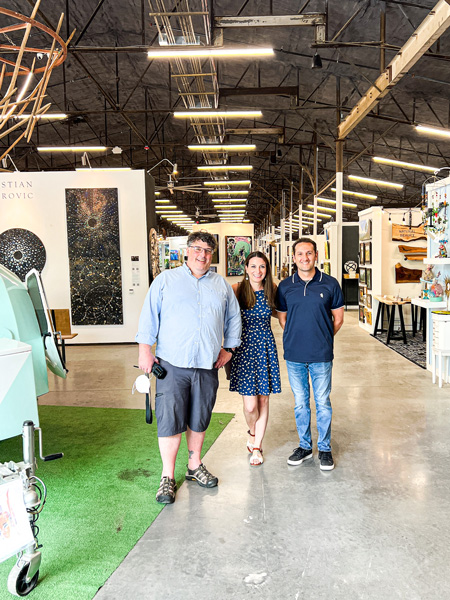 Marquee Asheville Shopping Center with two white men and brunette white female standing in large indoor art space filled with stalls