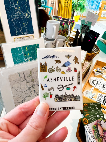 Moonlight Makers Asheville NC with white hand holding up magnet that says Asheville with icons of camping, Biltmore, mountains, and more