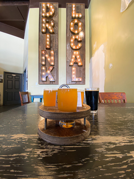 Oklawaha Brewing Company in Hendersonville NC Gluten Reduced Beer flight with three orange beers and dark stout on table with drink local lighted sign on the wall