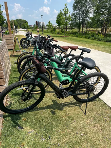 Reedy Rides Swamp Rabbit Trail Bike Rentals with photo of row of their bikes in various colors like black and green locked on bike rack on grass in front of brewery along the trail