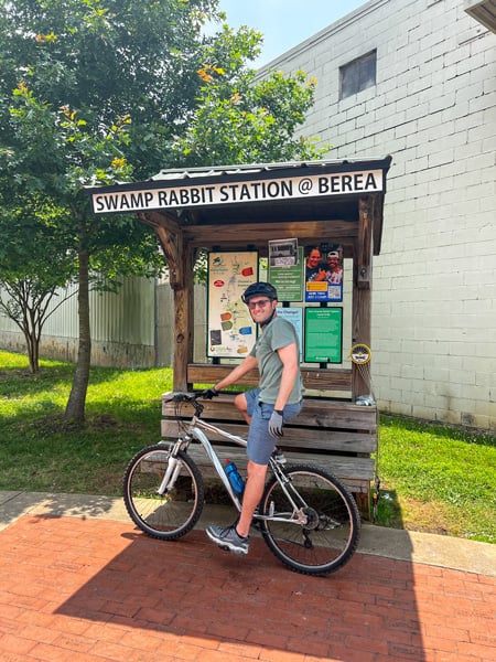 Swamp Rabbit Station At Berea with white male in helmet with sunglasses on mountain bike in front of trail information board with Swamp Rabbit Trail map