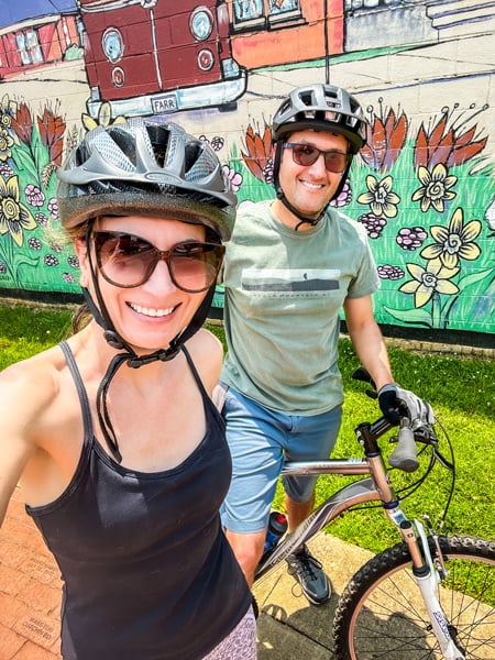 Christine and Tom, a white brunette male and female on mountain bikes with helmets and sunglasses, taking a selfie  in front of mural with flowers and grass at the Swamp Rabbit Station at Berea along the Swamp Rabbit Trail in Greenville, SC