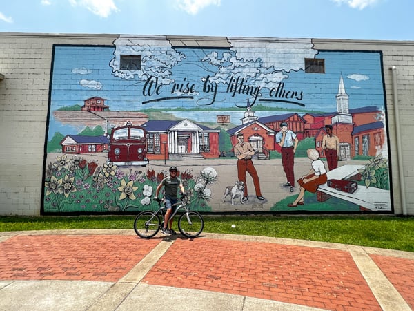 Swamp Rabbit Station at Berea Mural with town scape including four people, flowers, and building with quote that says we rise by lifting others with white brunette male on mountain bike in front of it