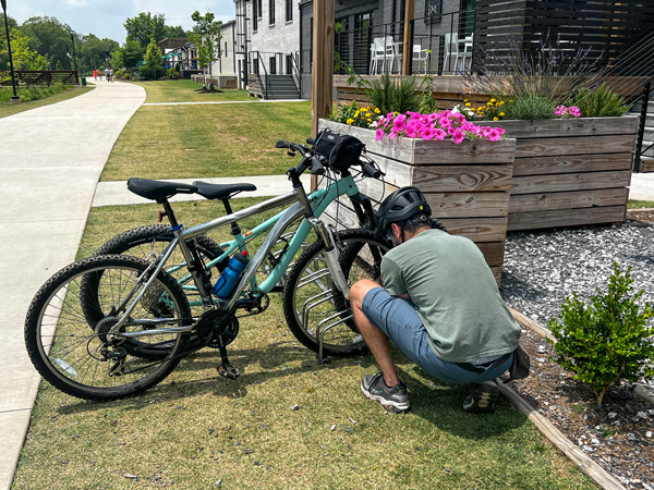 Swamp Rabbit Trail bike rack with male in blue shorts and green tshirt locking two bikes to rack along the trail