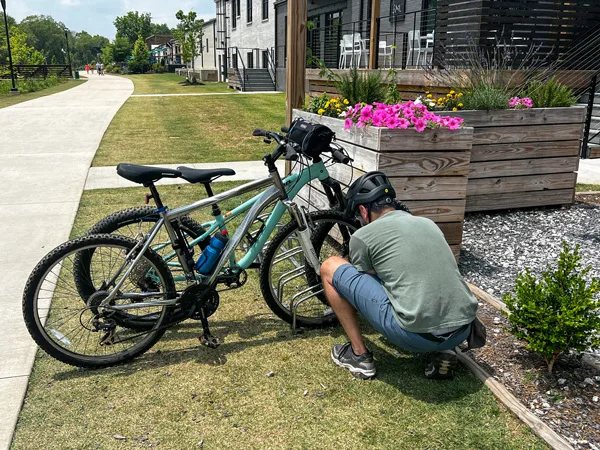 Swamp Rabbit Trail bike rack with male in blue shorts and green tshirt locking two bikes to rack along the trail