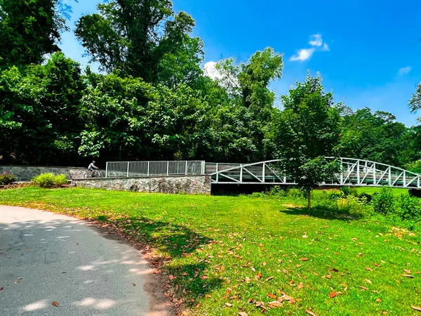 Swamp Rabbit Trail Cleveland Park Bridge in Greenville SC with mountain biker in distance going over steel bridge with green trees, green grass, and blue sky