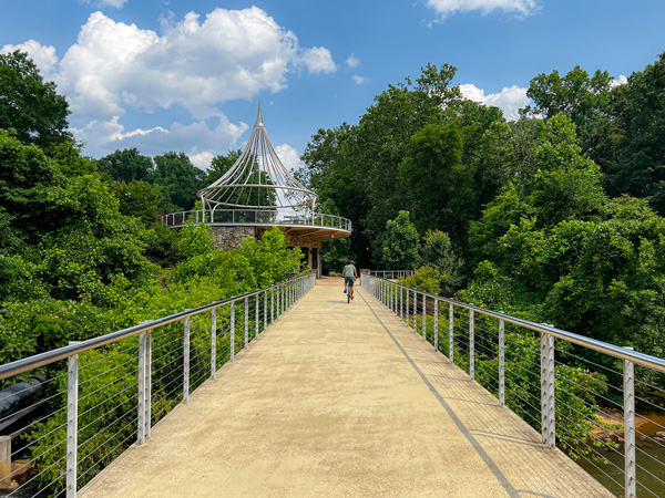 Swamp Rabbit Trail Greenville SC Park Bridge with mountain biker going across it in the distance and pavilion up ahead surrounded by green trees with blue cloudy sky