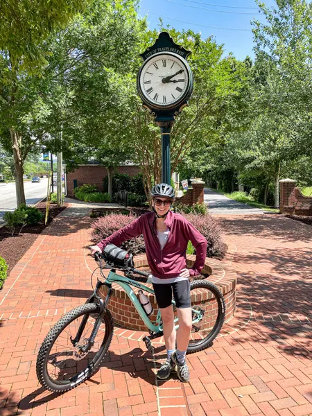 Swamp Rabbit Trail in Travelers Rest, SC Downtown clock with Christine, a white female in black biking shorts and pink jacket on deep mint green mountain bike with trees in the background