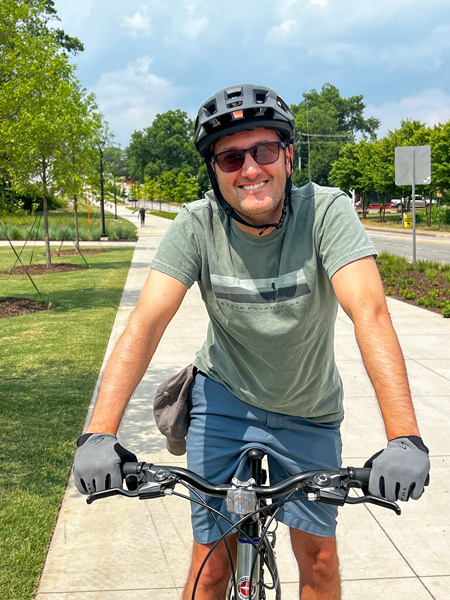 Tom, a white brunette male with helmet, sunglasses, gloves, and green t-shirt with blue shorts on mountain bike, along the Swamp Rabbit Trail at Unity Park in Greenville, SC