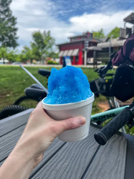 Whistle Stop at the American Cafe in Travelers Rest, SC Blue Shaved Ice held up by white hand outside in front of restaurant with bikes and Swamp Rabbit Trail in the background