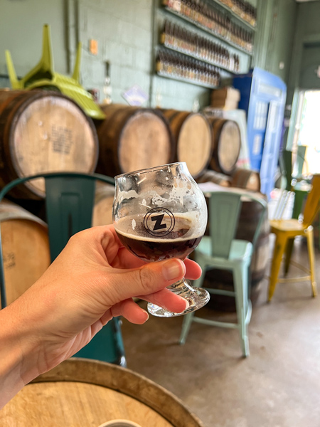 Zebulon Brewing Weaverville, NC brewery with white hand holding up brown ale in glass in front of barrels and green chairs in taproom