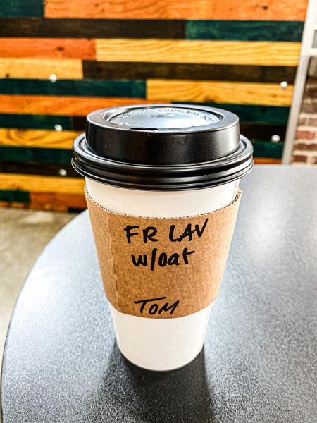 Bebettes Coffee Downtown Asheville NC with white to-go cup that says FR LAV with oat and Tom sitting on table with shiplap wall in background