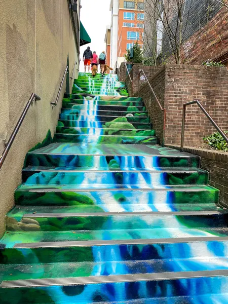 Downtown Asheville Waterfall Mural along concrete staircase with white, blue, and green coloring, and people walking up them at the top