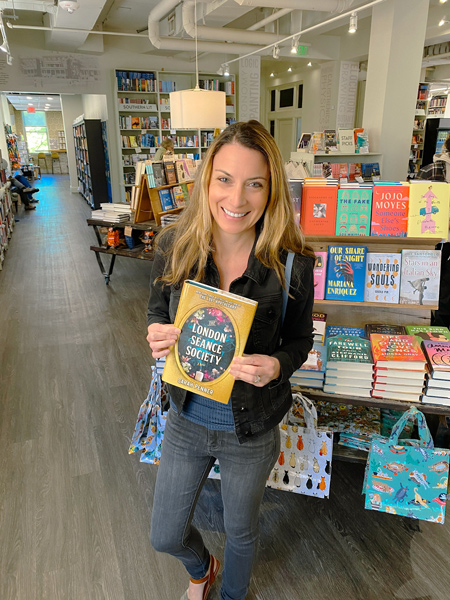 M Judson Booksellers in Greenville SC with white brunette woman with blonde highlights holding up a book in bookshop in front of shelves