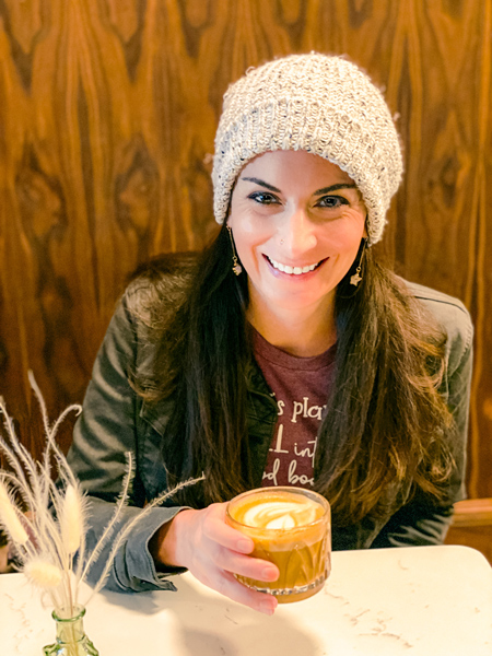 Christine, a white brunette female in a white hat with green jacket and purple shirt drinking a latte at a table at Rowan Coffee in Downtown Asheville, NC