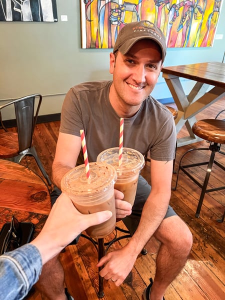 The Rhu Coffee in Downtown Asheville with white brunette male in hat and t-shirt doing a cheers with another white hand holding iced to-go coffee