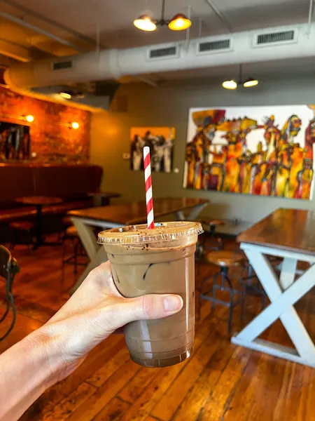 The Rhu Coffee Shop in Downtown Asheville Upstairs seating area with paintings, tables, and white hand holding up to-go ice coffee with red and white striped paper straw