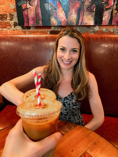 The Rhu Downtown Asheville Coffee Shop with white brunette woman with blonde highlights in a dress doing a cheers with iced coffee