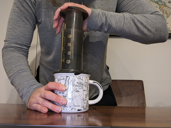 Aeropress Coffee Press with white hand and person in gray top pressing coffee over white mug