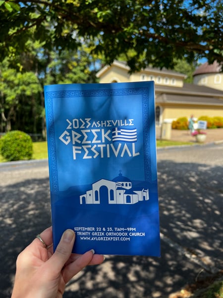 Asheville Greek Festival 2023 Program, blue with white writing, held up in front of the Holy Trinity Greek Orthodox Church of Asheville